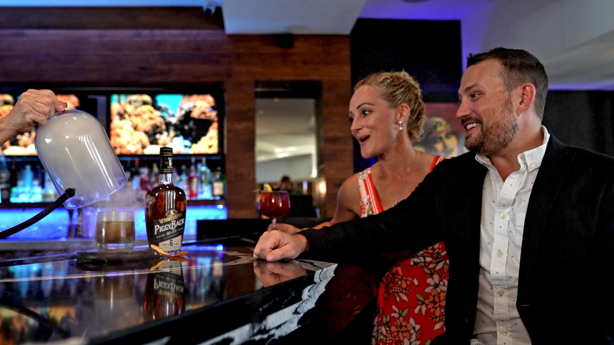 Two people sitting at a bar with drinks, watching a bartender create a smoky effect with a glass dome. A bottle of Pendleton Whiskey is on the counter.