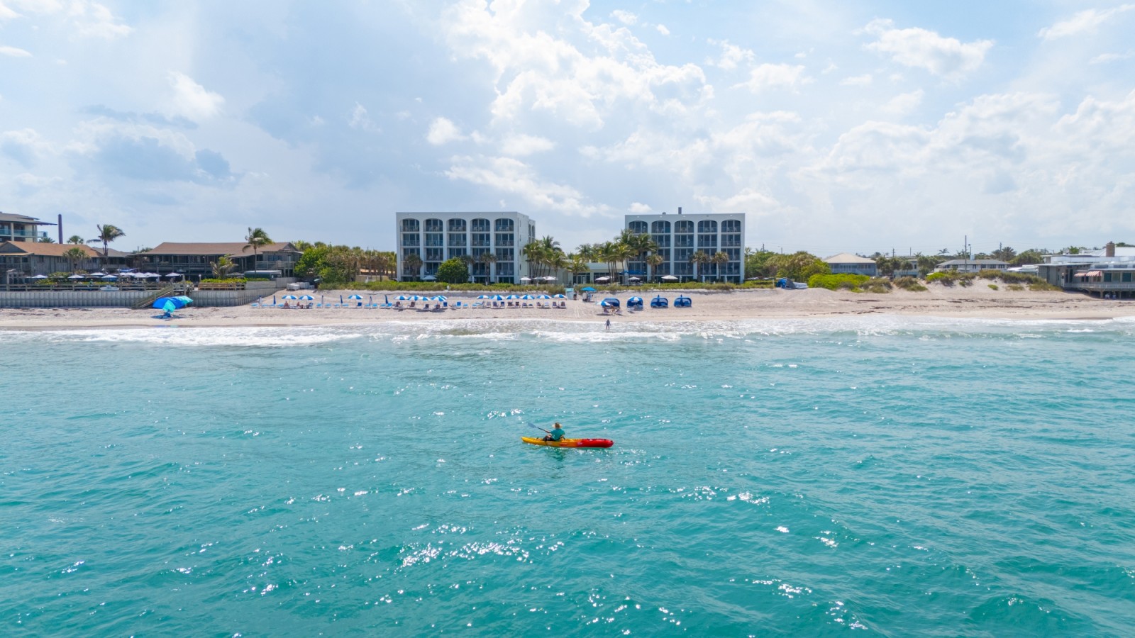 A sunny beach scene with turquoise water, a few people on the shore, and two modern buildings in the background; a small boat floats near shore.