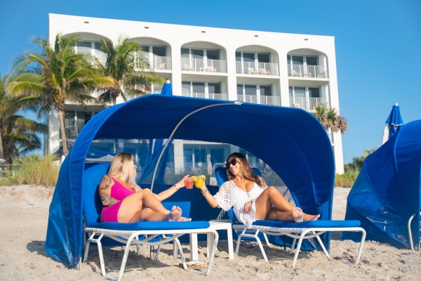 Two women lounging under a blue beach cabana on the sand, clinking drinks with a resort building behind.