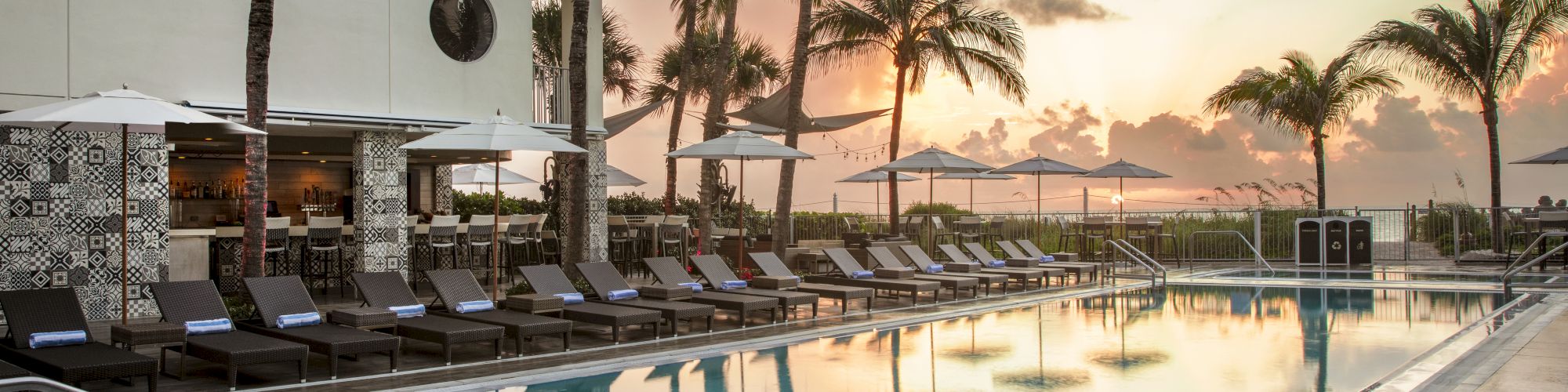 A tranquil outdoor pool area with lounge chairs, umbrellas, and palm trees at sunset; a perfect spot for relaxation and enjoyment.