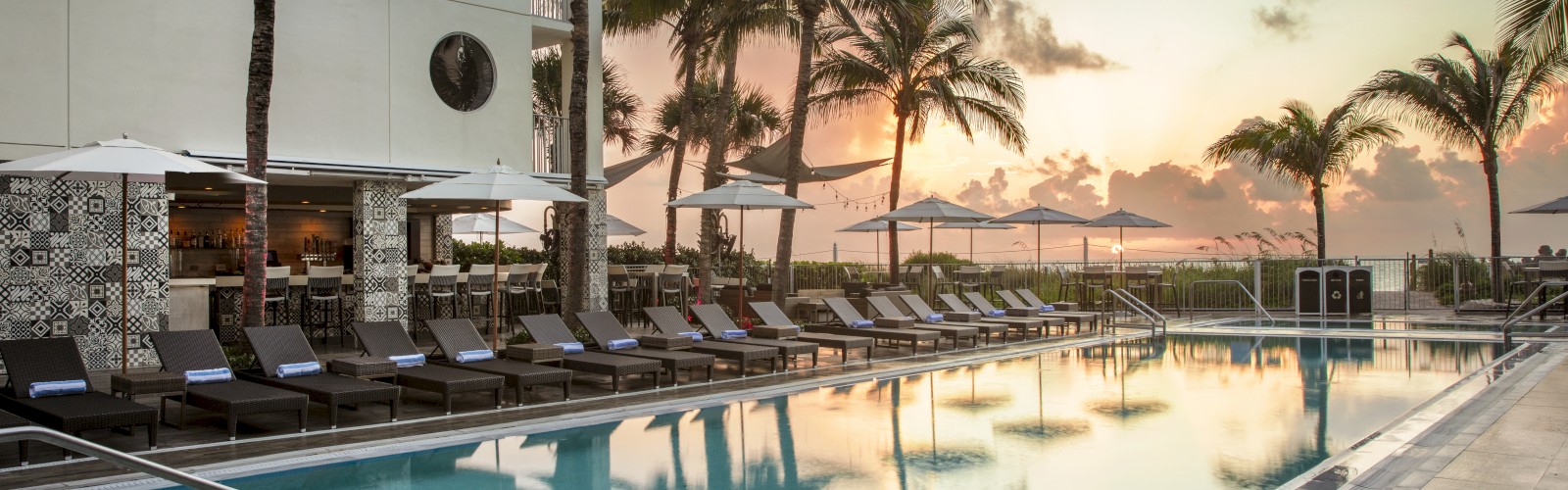 A tranquil outdoor pool area with lounge chairs, umbrellas, and palm trees at sunset; a perfect spot for relaxation and enjoyment.