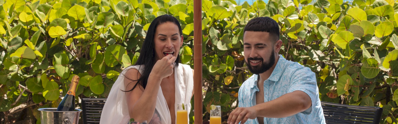 A couple enjoys a meal outdoors at a table shaded by an umbrella, with lush greenery and the ocean in the background.