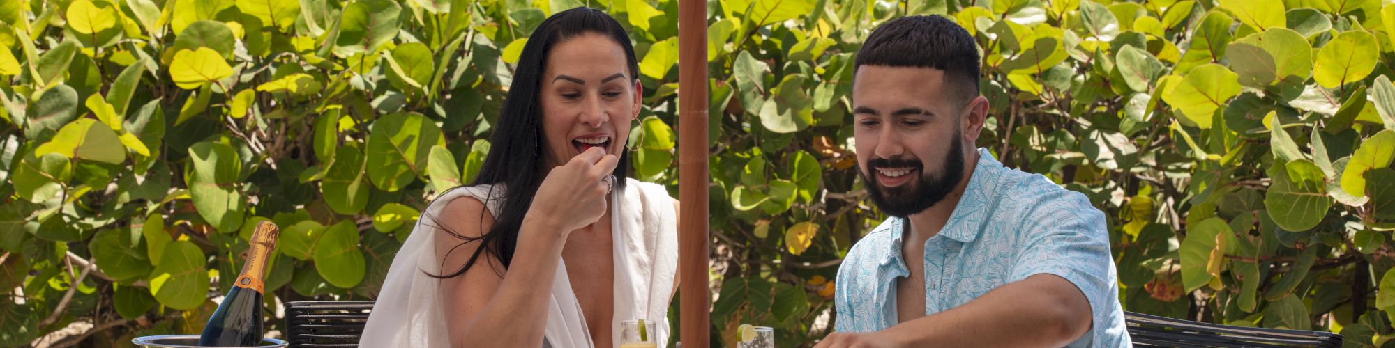 A couple enjoys a meal outdoors at a table shaded by an umbrella, with lush greenery and the ocean in the background.