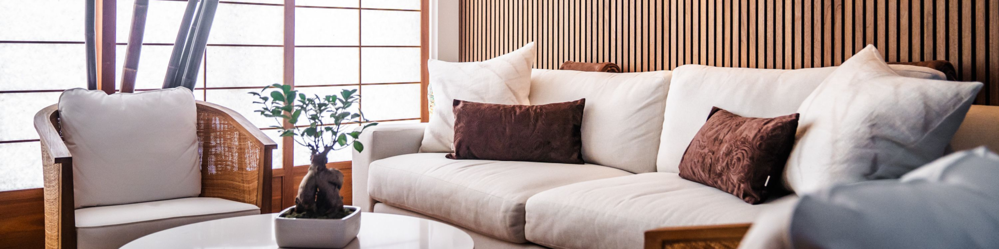 A modern living room with white furniture, brown cushions, wooden panel walls, warm lighting, and a small table with a plant in the center.