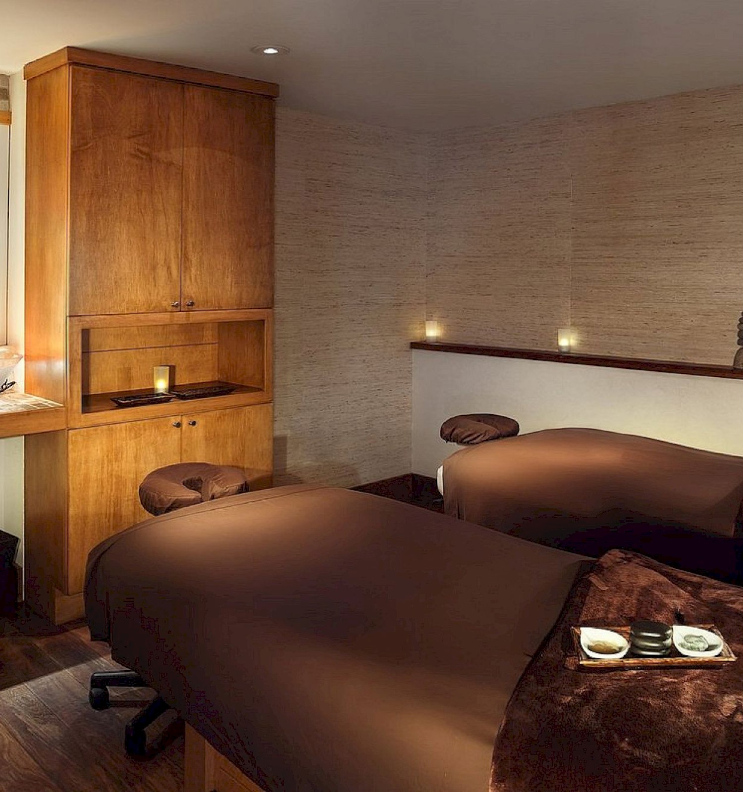A cozy spa room with two massage tables, dim lighting, warm wooden accents, candles, and a sink area for relaxation and treatments.