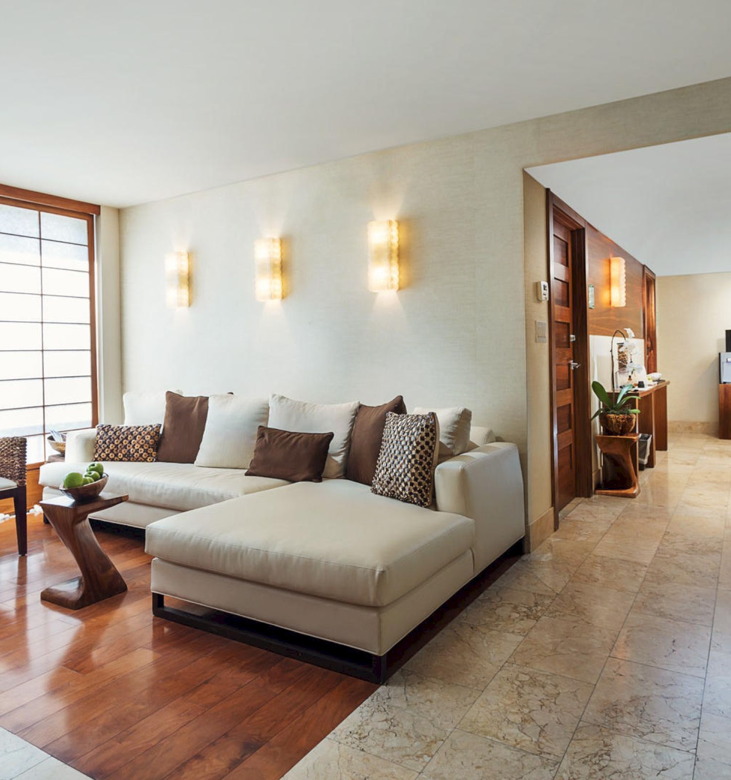 A modern living room features a white sectional sofa, a wooden coffee table, woven chairs, and ambient wall lights, leading to a hallway.