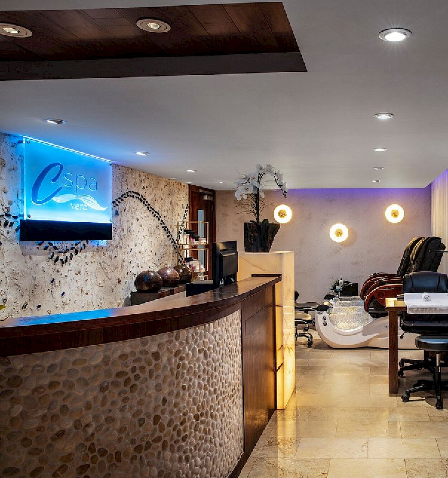 The image shows a modern spa reception area with a massage chair, shelves stocked with products, and a curved counter.