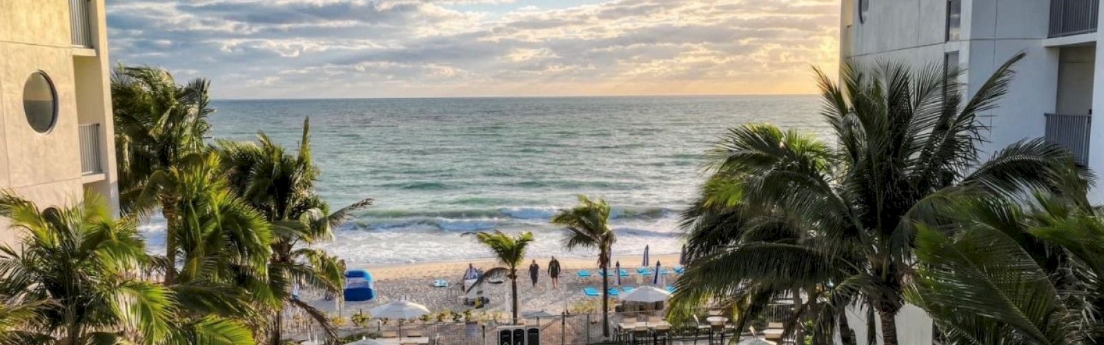A scenic view of a resort features a pool surrounded by lounge chairs and umbrellas, with a backdrop of a beach and ocean waves under a partly cloudy sky.