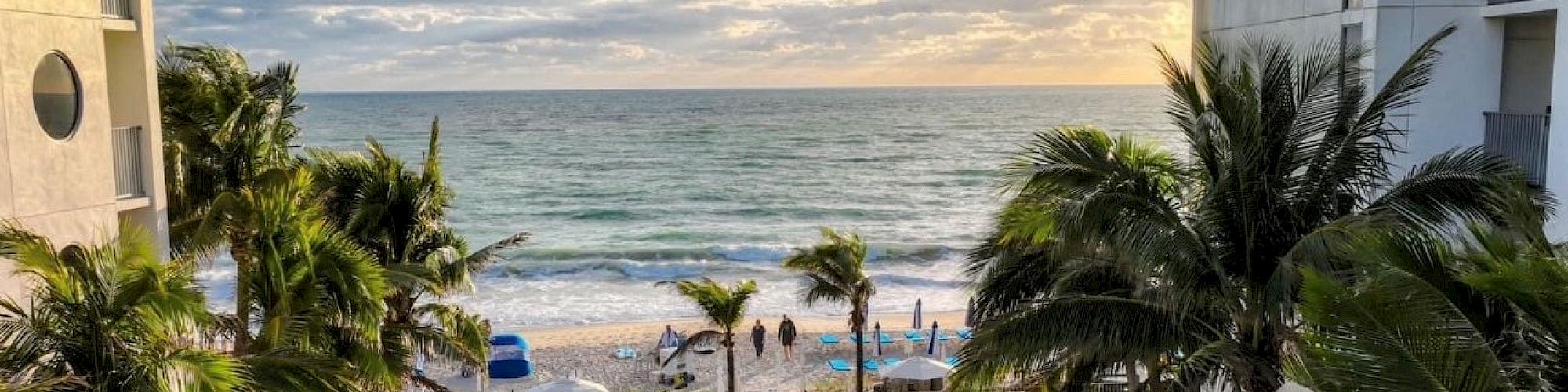 A scenic view of a resort features a pool surrounded by lounge chairs and umbrellas, with a backdrop of a beach and ocean waves under a partly cloudy sky.