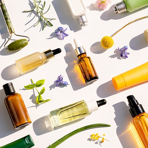 The image shows various skincare and beauty product bottles and tubes among flowers and greenery on a white background.