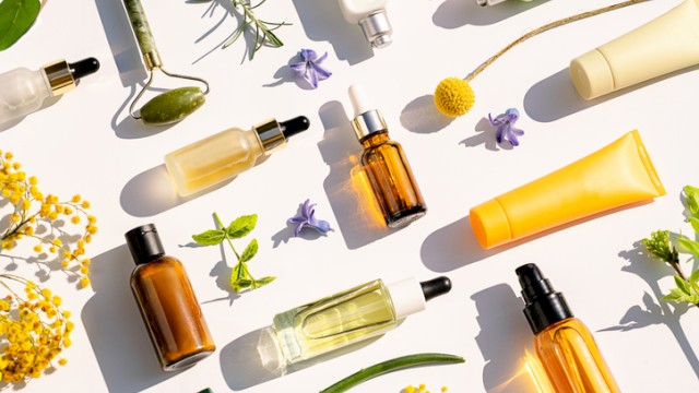 The image shows various skincare and beauty product bottles and tubes among flowers and greenery on a white background.