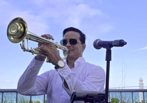 A man in sunglasses plays a trumpet outdoors next to a microphone, with a blue sky and greenery in the background.