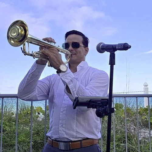 A man in sunglasses plays a trumpet outdoors next to a microphone, with a blue sky and greenery in the background.