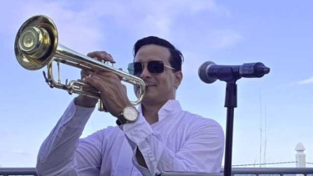 A man in sunglasses plays a trumpet outdoors next to a microphone, with a blue sky and greenery in the background.