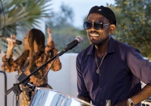 A man wearing sunglasses and a cap plays a shiny steel percussion drum on a sunny outdoor stage, with a female singer in the background.