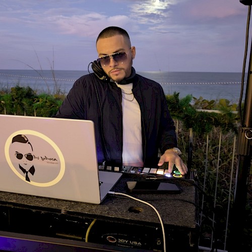A DJ wearing sunglasses and a black jacket, mixing music on a laptop and turntables by the beach at sunset. End with a period.