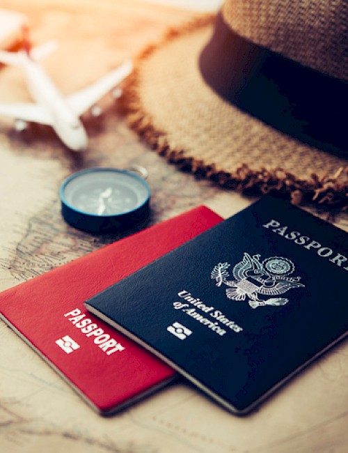The image shows two passports, a red one and a navy blue one, along with a small toy airplane, a cork hat, a notebook, and a pencil on a travel map.