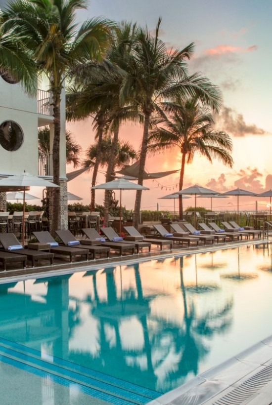A luxurious poolside area with sun loungers, umbrellas, palm trees, and a beautiful sunset sky in a tropical setting.