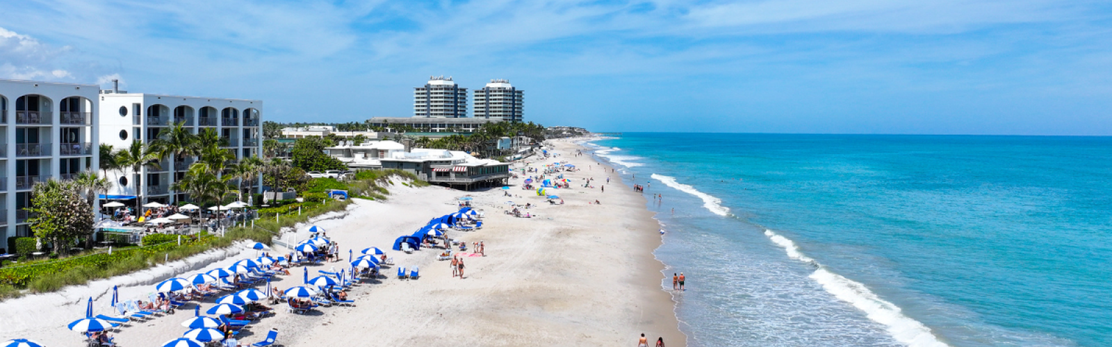 A sunny beach scene with umbrellas, lounge chairs, and people enjoying the ocean and sand under a blue sky.
