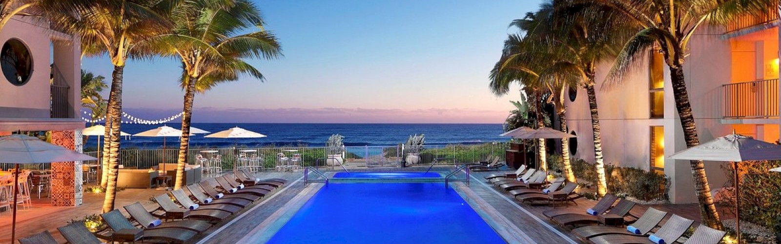 A luxury poolside area with lounge chairs and palm trees, overlooking a serene beach and ocean during sunset, creating a tranquil atmosphere.