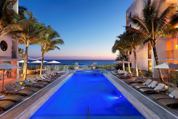 A luxurious outdoor pool area with lounge chairs, surrounded by modern buildings and palm trees, overlooking a beautiful ocean sunset.