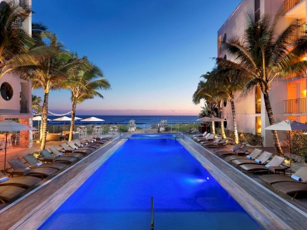 A luxurious outdoor pool area with lounge chairs, surrounded by modern buildings and palm trees, overlooking a beautiful ocean sunset.