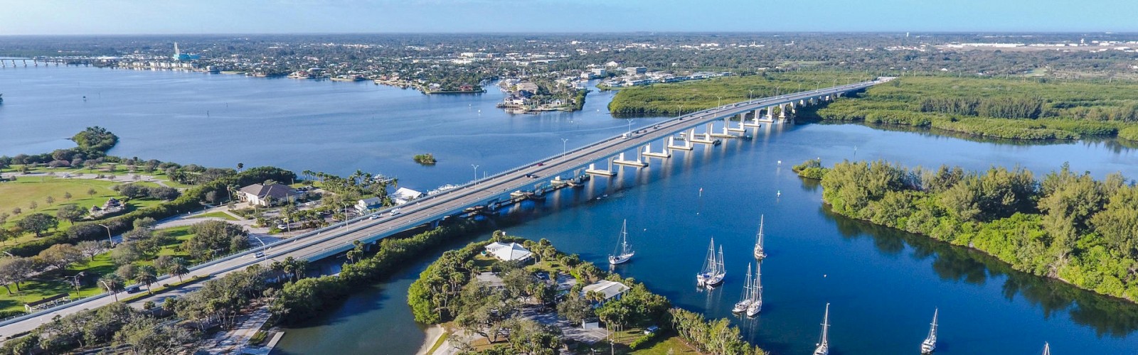 This image shows a scenic view of a coastal area with a bridge over water, boats, green landscapes, and a distant town in the background.