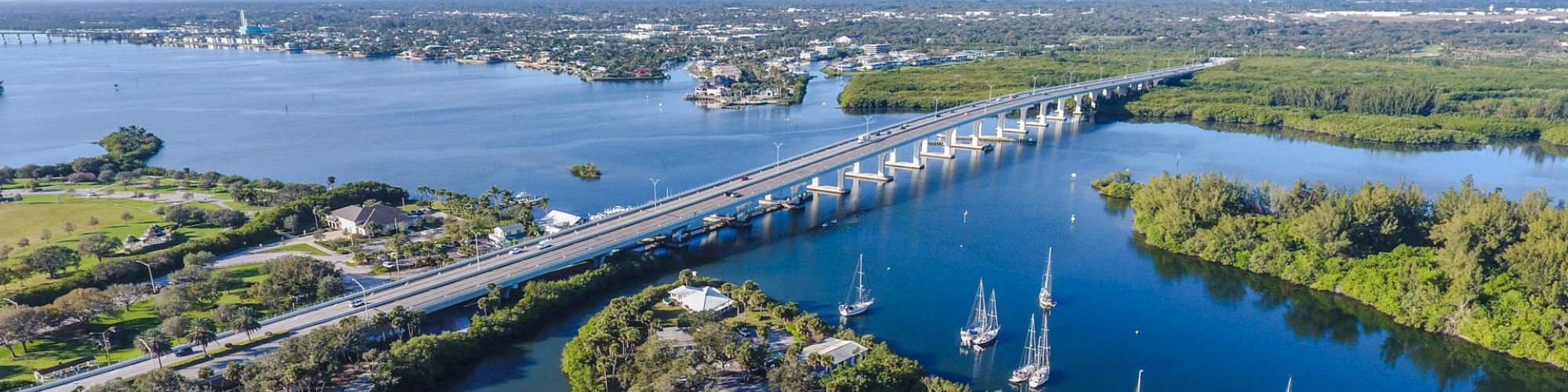 This image shows a scenic view of a coastal area with a bridge over water, boats, green landscapes, and a distant town in the background.