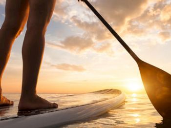 A person paddleboarding at sunset, their legs on the board and paddle in hand. The sun sets over calm waters, creating a serene scene.