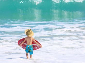 A child wearing a hat and swim trunks holds a boogie board while standing in shallow ocean water, ready to catch some waves.