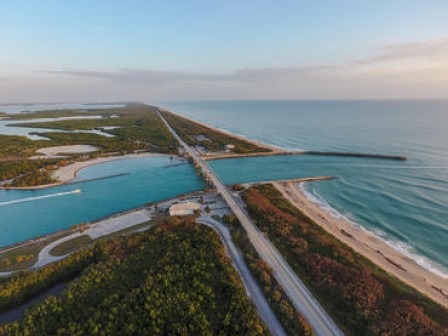 Aerial view of a bridge spanning a waterway, with lush green areas and the ocean on either side, leading to a long coastal road.