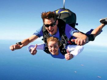 Two people are tandem skydiving over a large body of water with the horizon in the background; both appear to be enjoying the experience.