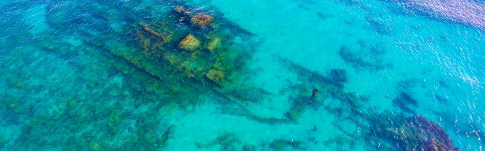 The image shows a sunken shipwreck in clear blue-green shallow water. The outline of the ship is visible beneath the surface.