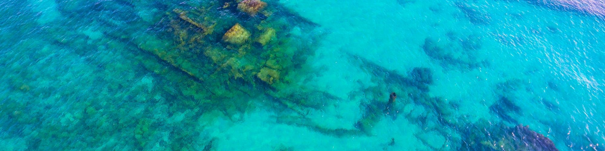The image shows a sunken shipwreck in clear blue-green shallow water. The outline of the ship is visible beneath the surface.