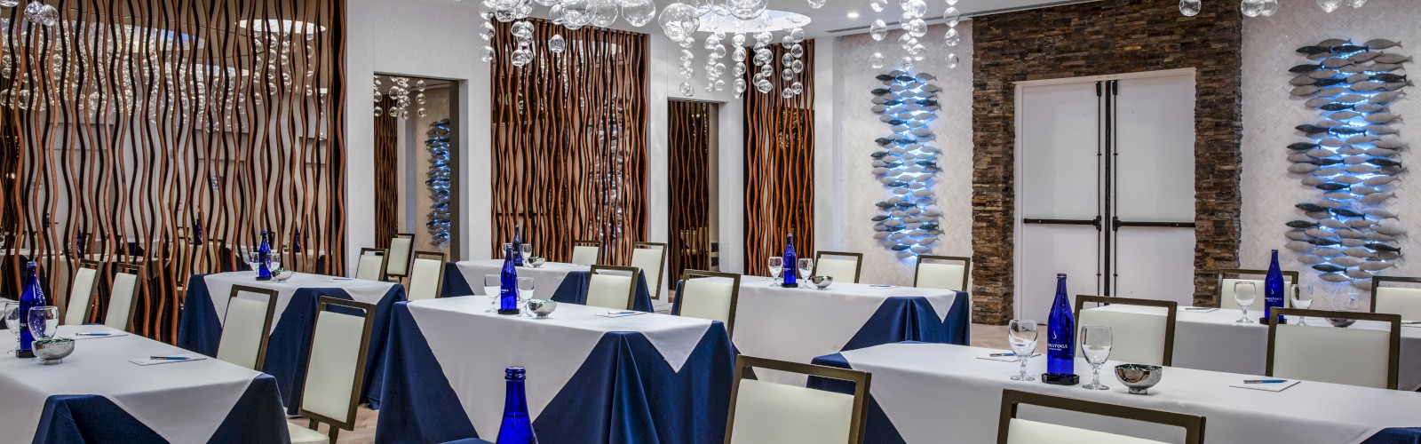 A modern conference room with tables set with blue bottles and glasses, elegant lighting fixtures, and white walls adorned with artistic decor.