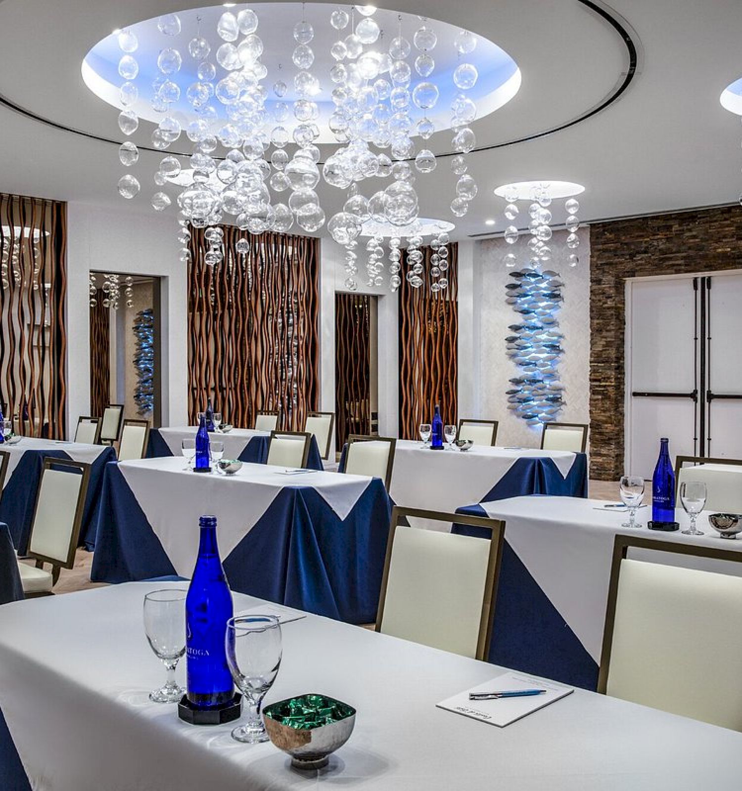 A conference room with tables set up, blue bottles, glasses, notepads on tables, and modern hanging lights from the ceiling, ending the sentence.