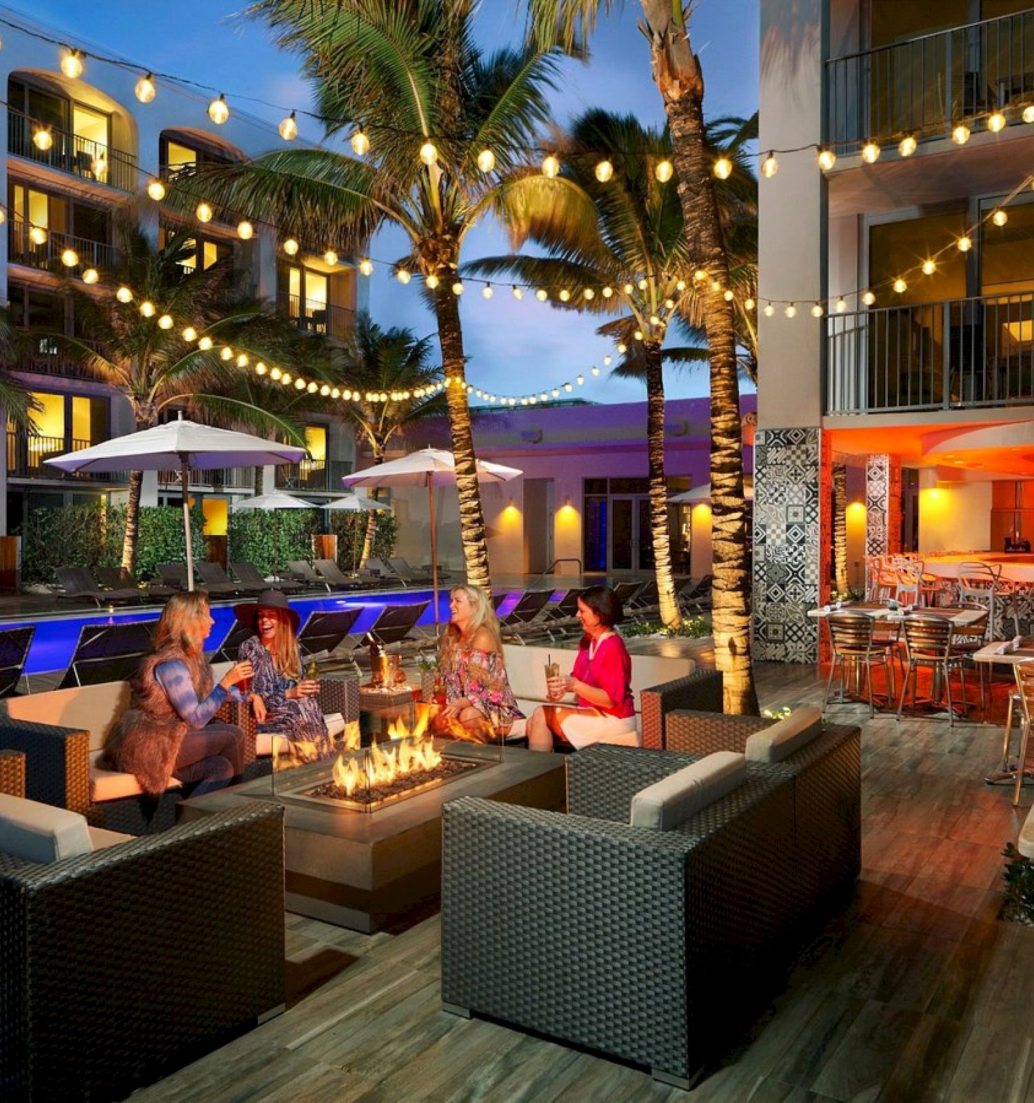People are gathered around a fire pit in an outdoor lounge area of a resort, with string lights, palm trees, and a pool in the background.