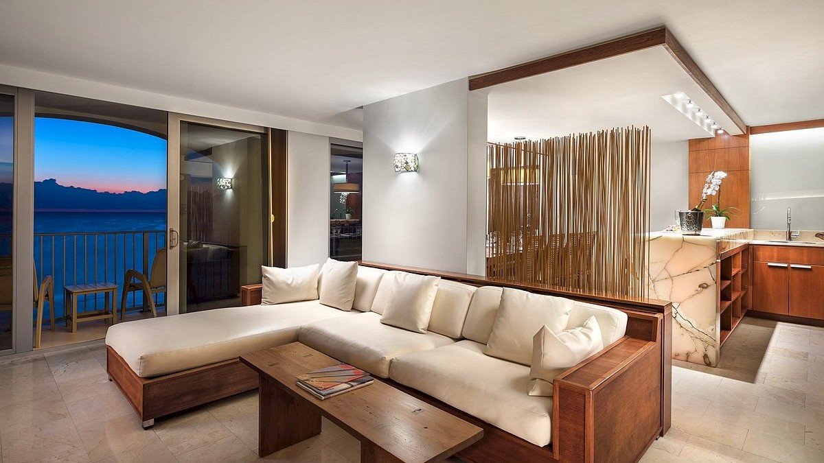 A modern living space featuring a white sectional sofa, a wooden coffee table, a kitchen area with a marble countertop, and a balcony view of the ocean.