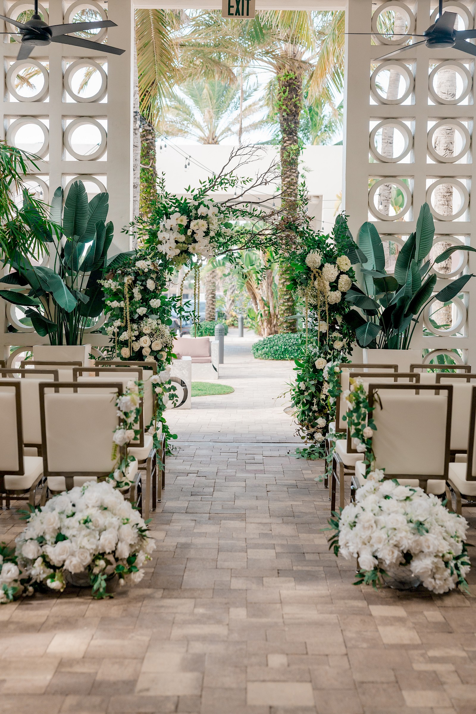 A beautifully decorated wedding aisle with white flowers and greenery, set outdoors under a canopy with tropical foliage in the background.