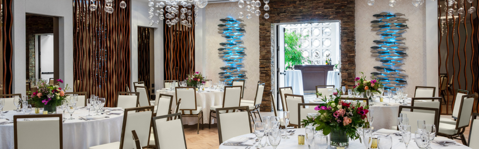 A beautifully decorated event space with round tables set for dining, elegant chandeliers, floral centerpieces, and modern wall art and design elements.