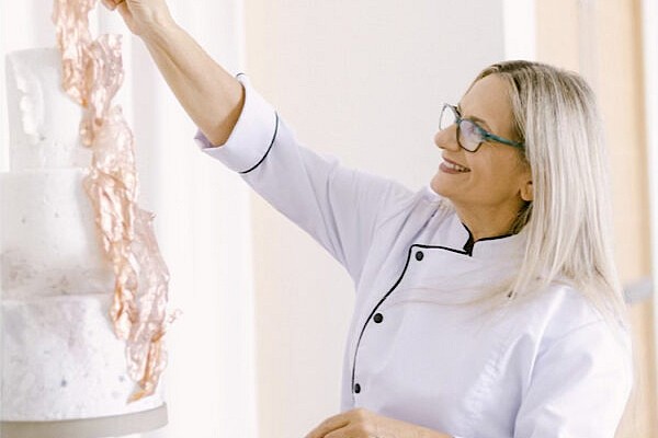 A person in a chef's coat is decorating a two-tier cake with pink ribbons, smiling and focused on their work in a bright room.