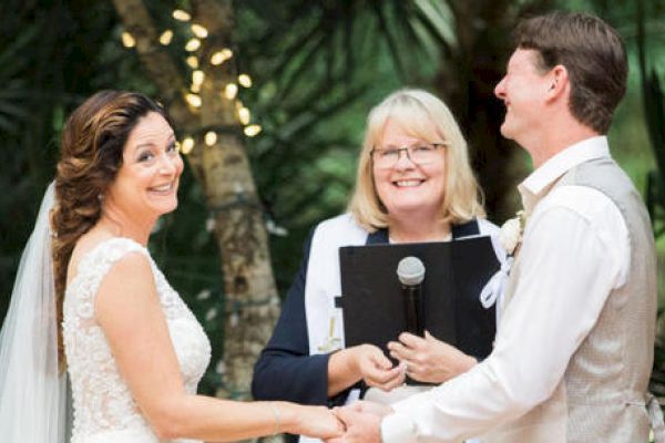 A bride and groom are holding hands during a wedding ceremony, with a smiling officiant standing between them.