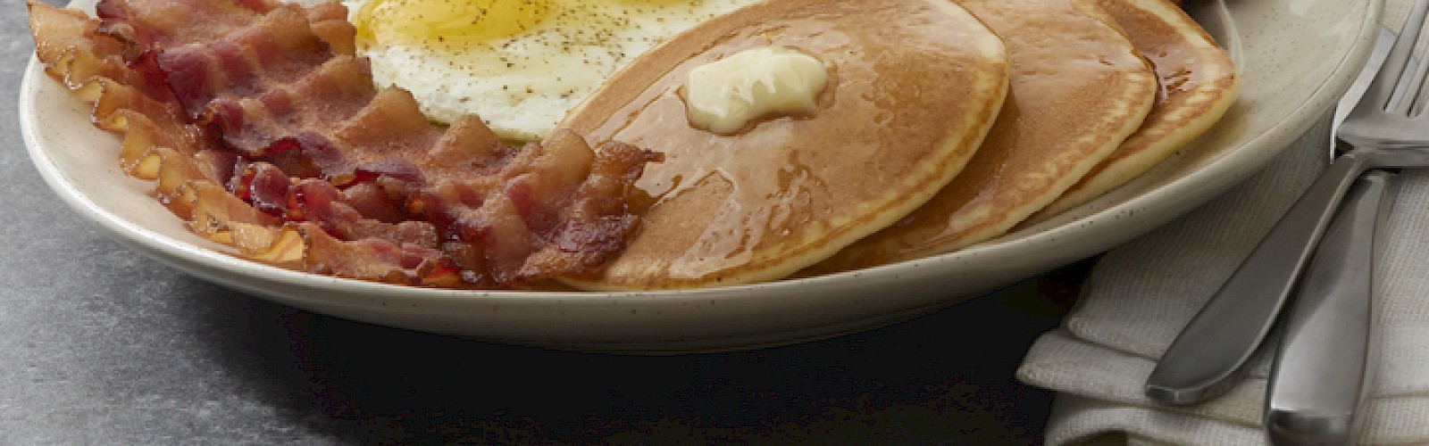 A traditional breakfast with fried eggs, bacon, pancakes, sausages, toast, orange juice, and coffee on a plate.