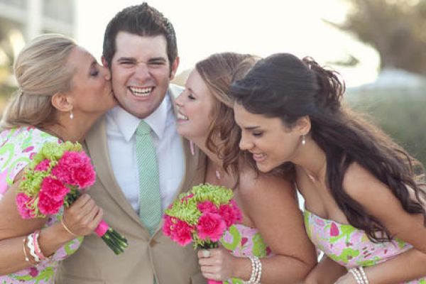 A man in a tan suit is happily posing with three women holding bouquets, showing affection towards him in a cheerful outdoor setting.