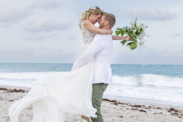 A couple is embracing on a beach; the woman holds a bouquet and wears a flowing dress, while the man lifts her in a joyful pose.