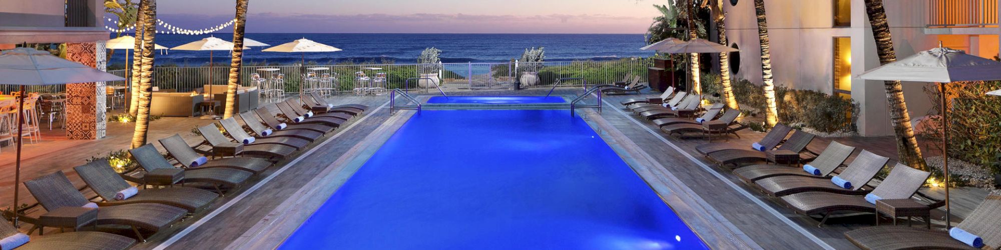 An illuminated swimming pool surrounded by lounge chairs and palm trees at a resort, with an oceanside view at sunset.