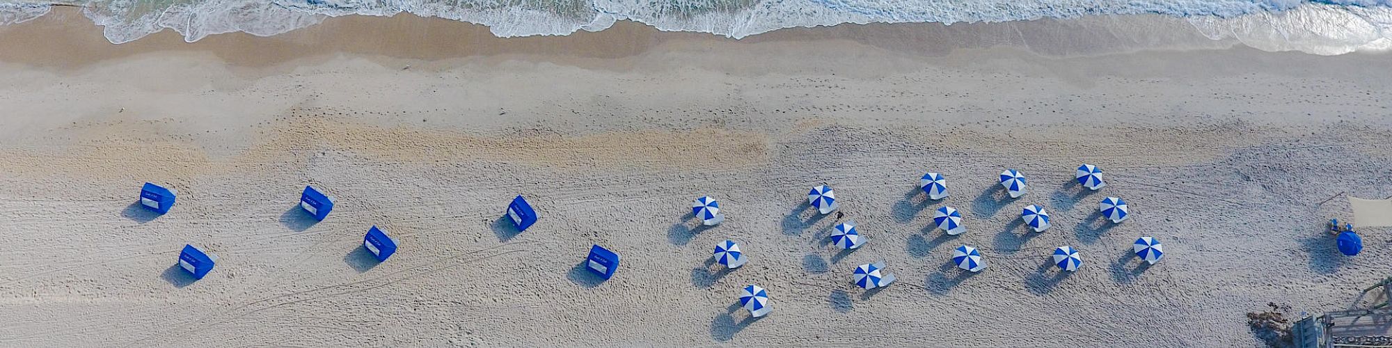 An aerial view of a beach with lined-up blue and white umbrellas, beach chairs, and the ocean's waves touching the shore in the background.