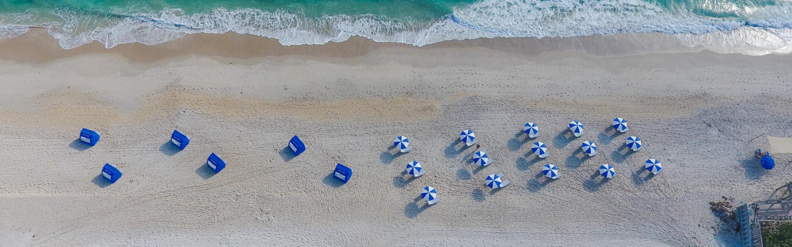 An aerial view of a beach with lined-up blue and white umbrellas, beach chairs, and the ocean's waves touching the shore in the background.