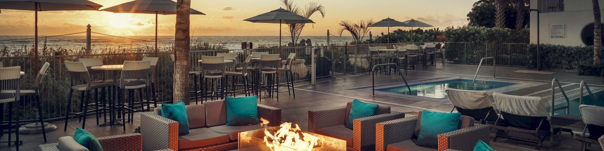 Outdoor seating area with wicker furniture, cushioned chairs, fire pit, string lights, and pool against a sunset beach backdrop.