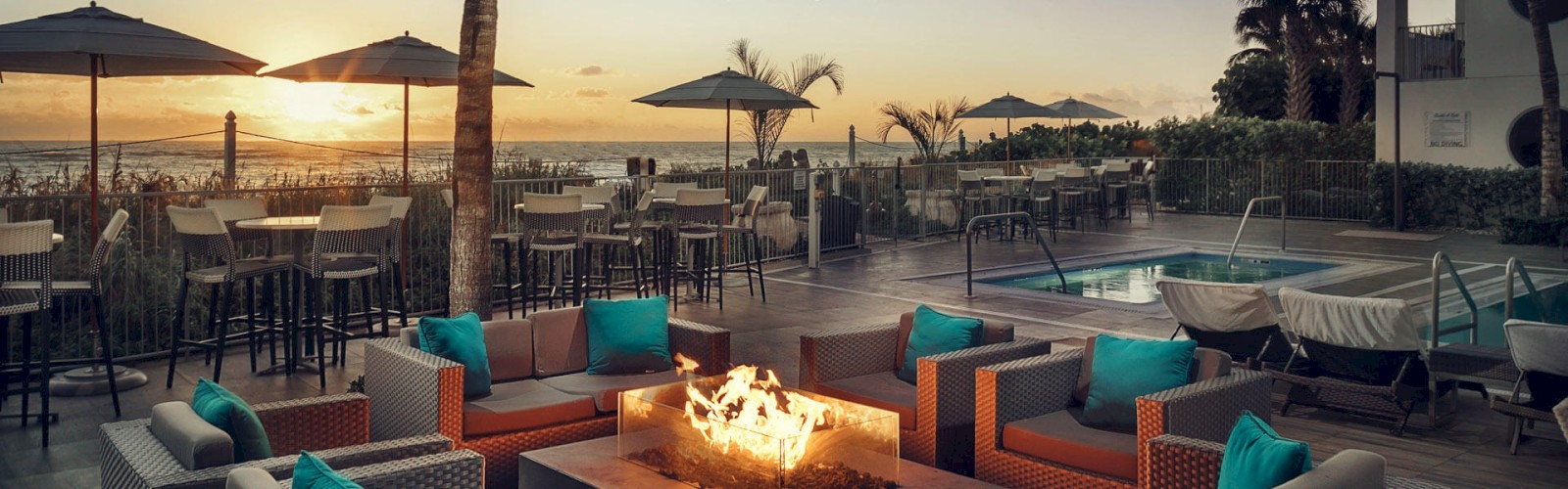 Outdoor seating area with wicker furniture, cushioned chairs, fire pit, string lights, and pool against a sunset beach backdrop.